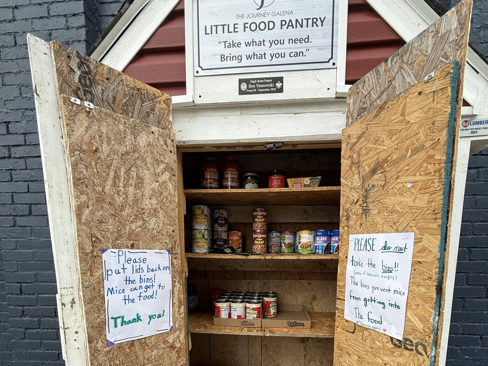 The Little Food Pantry cabinet outside the building, doors open and shelves stocked with canned goods. The sign reads: take what you need, bring what you can.